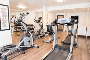 Un gimnasio con diversas máquinas de cardio, incluyendo cintas de correr y bicicletas estáticas, dispuesto en una sala amplia con piso de madera.