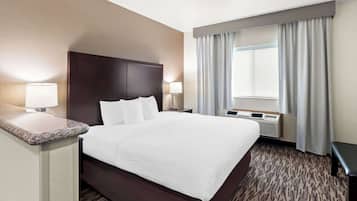 A hotel room with a large bed, two bedside tables with lamps, a window with curtains, and a wall-mounted air conditioning unit.