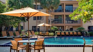 Zona de piscina con sillones de descanso y sombrillas, rodeada por un edificio con balcones.