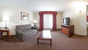 Habitación de hotel con sofá, mesa de centro, televisor en una cómoda, lámpara, un cuadro en la pared y ventana con cortinas.