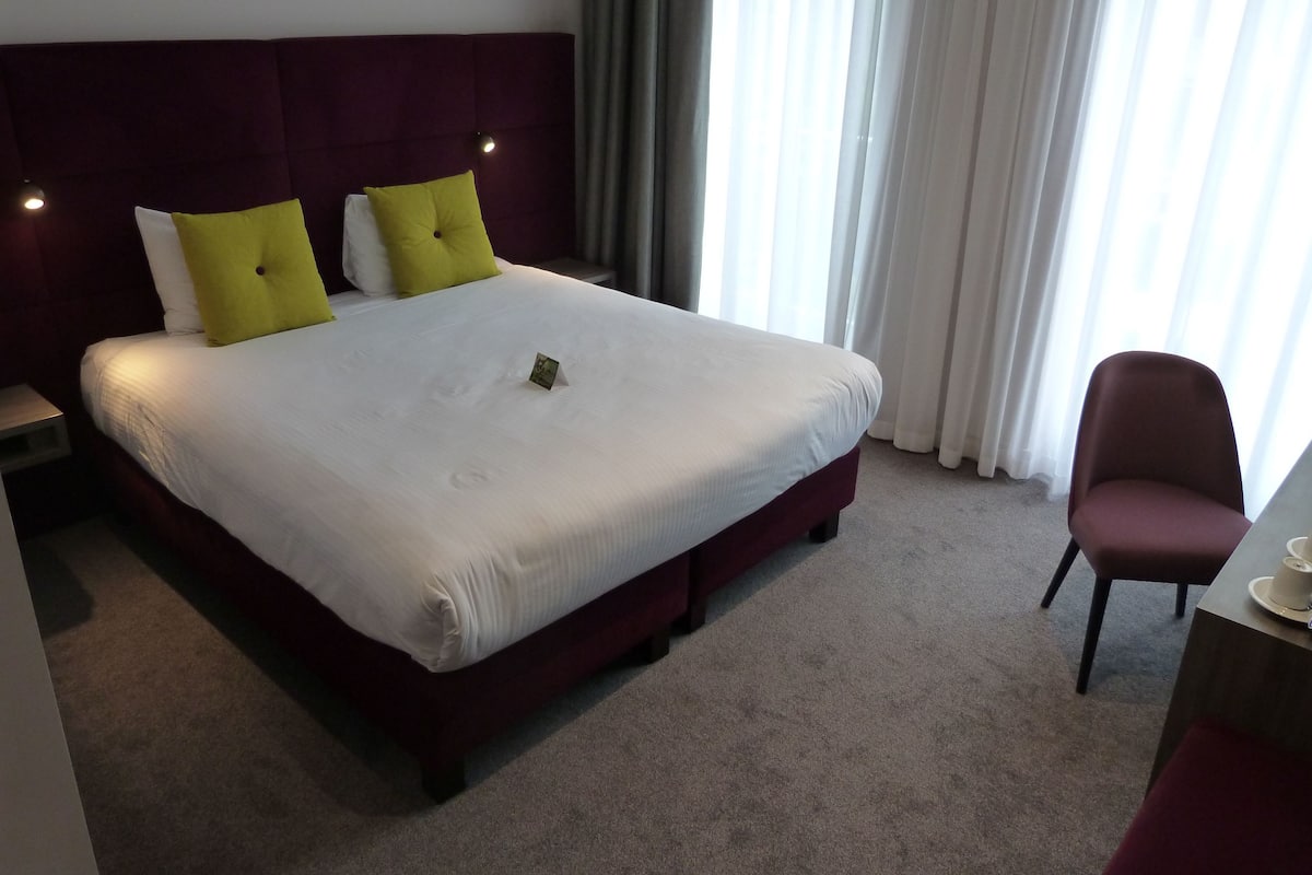 A hotel room with a large bed, two pillows, a bedside table, a chair, and a window with curtains.