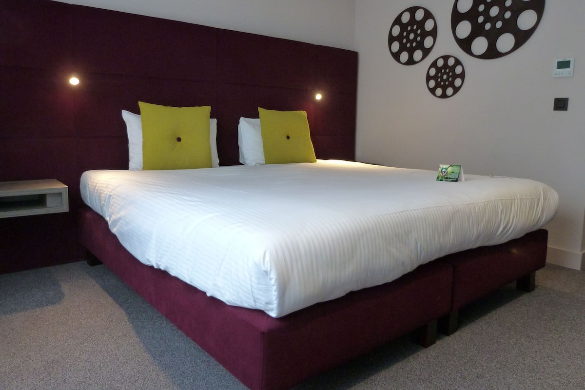 A large bed with white bedding, two yellow pillows, and a dark red headboard. There are circular wall decorations and a small green sign on the wall.
