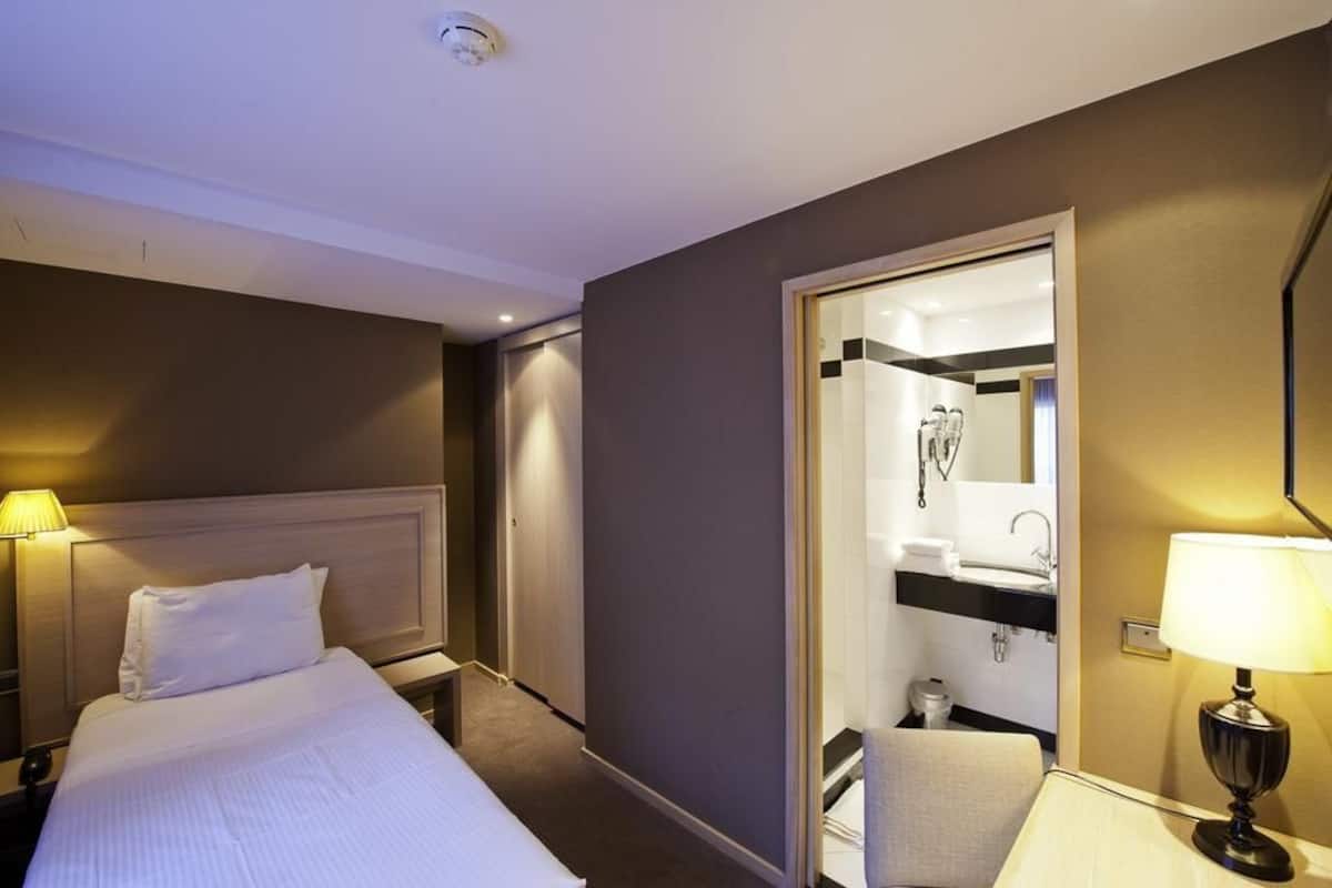 A hotel room with a bed, bedside lamps, a bathroom with a sink and mirror, and a door leading to another room.