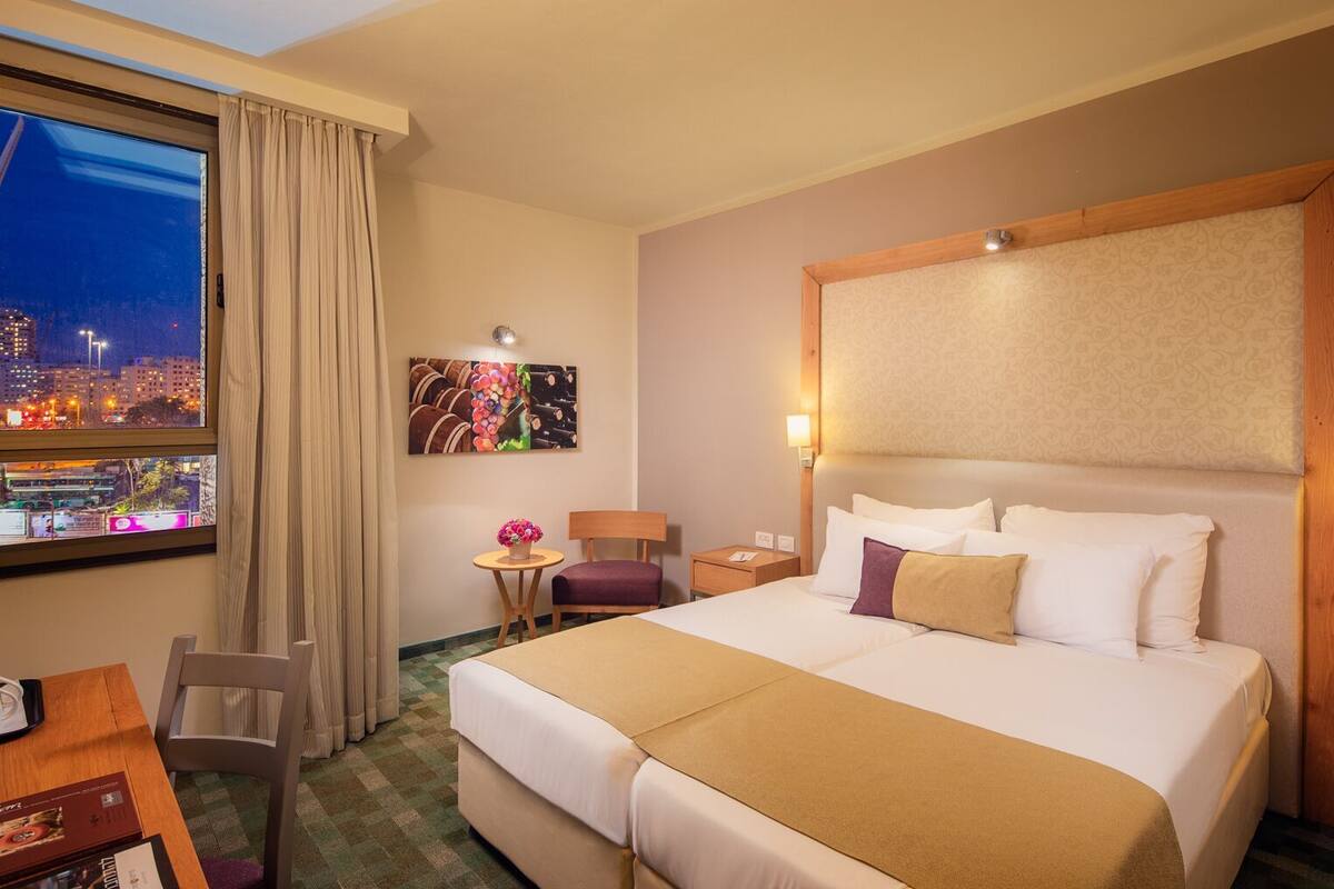 A hotel room with a large bed, a desk, a chair, a small table with a vase of flowers, and a city view through the window.