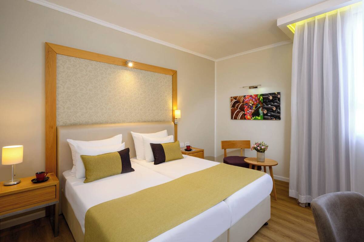A hotel room with a large bed, a bedside table with a lamp, a small table with a vase of flowers, and a chair.