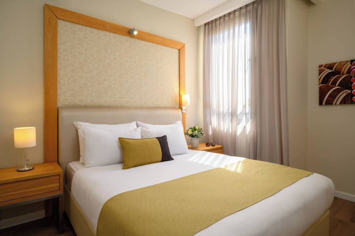A hotel room with a large bed, a wooden headboard, a bedside table with a lamp, and a window with curtains.