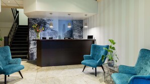 Una zona de recepción con un mostrador oscuro, dos sillones color turquesa, una mesita auxiliar con una planta y una escalera al fondo.