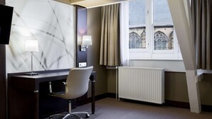 Habitación de hotel con escritorio, dos lámparas, una silla, radiador y ventana con cortinas.