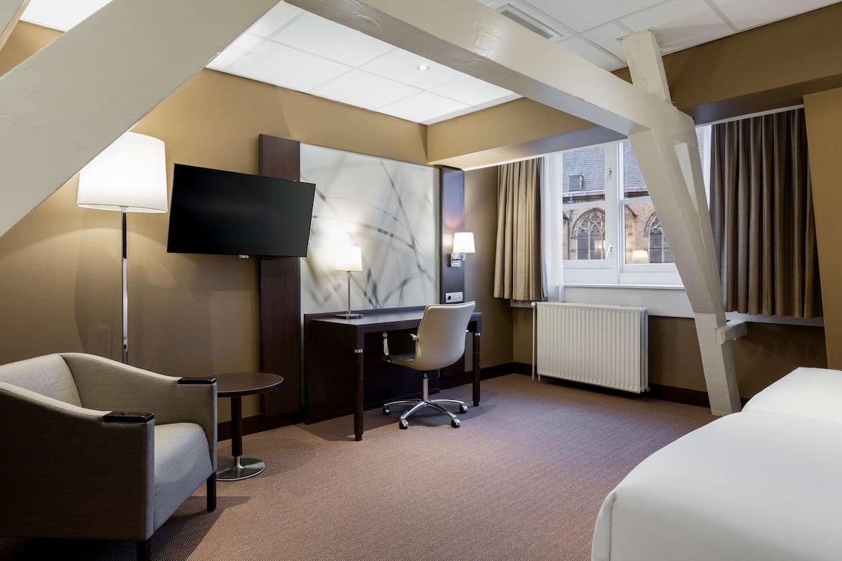 Habitación de hotel con cama, escritorio, silla, televisor y ventana con cortinas.