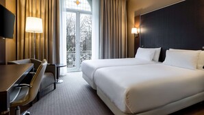 Habitación de hotel con una cama grande, un escritorio y una silla. Tiene una ventana con cortinas y vistas a árboles fuera.