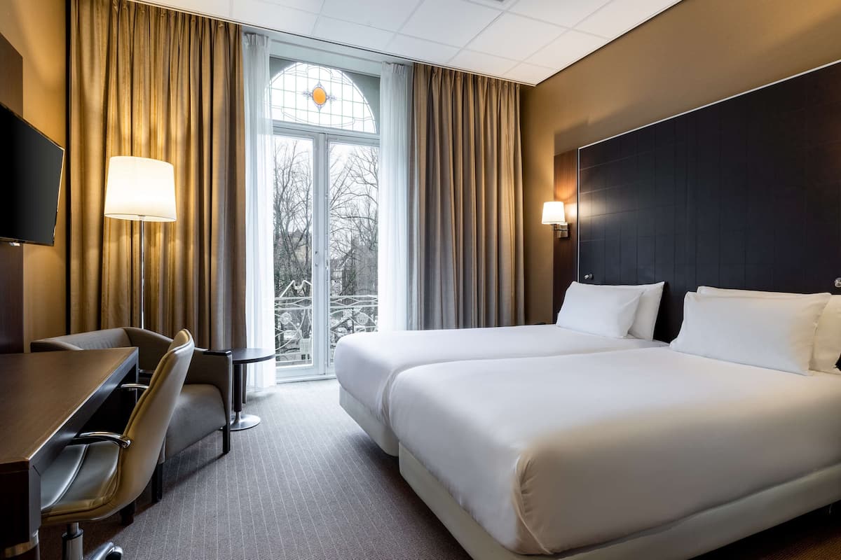 Habitación de hotel con una cama grande, un escritorio y una silla. Tiene una ventana con cortinas y vistas a árboles fuera.