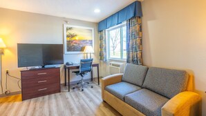 A room with a TV, desk, chair, sofa, and a window with curtains.