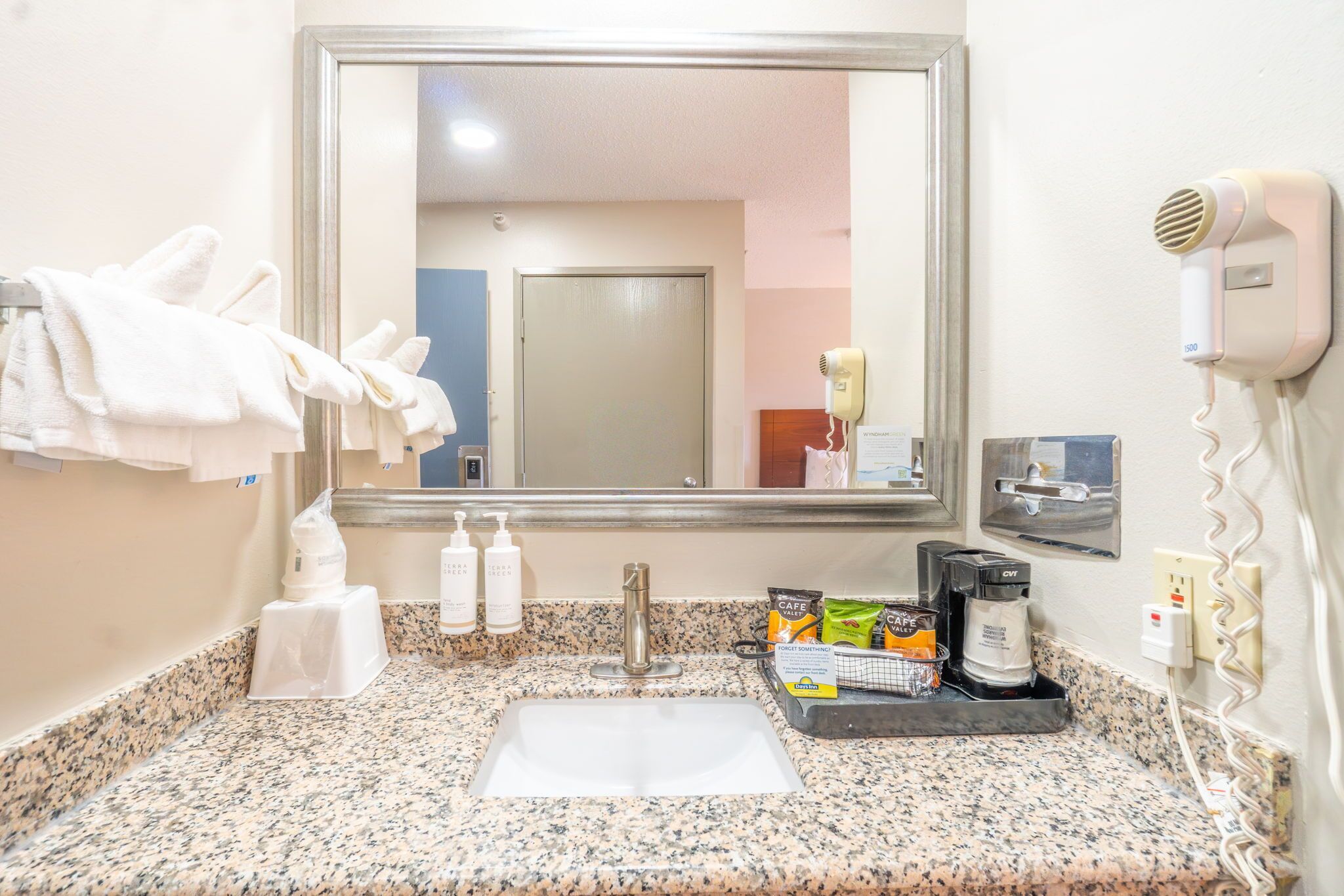 A hotel bathroom with a granite countertop, a sink, a mirror, a hairdryer, and toiletries.