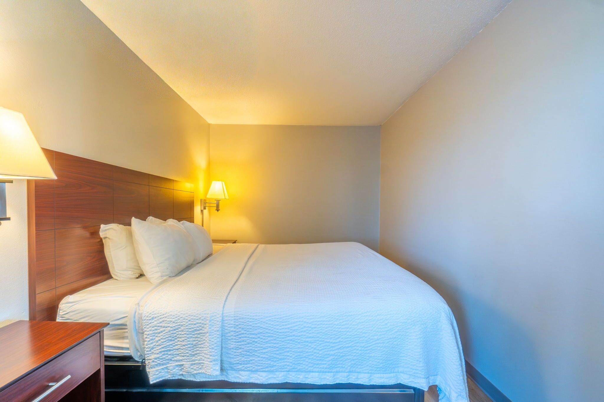 Habitación de hotel con cama, cabecero de madera, mesita de noche y lámpara de pared.