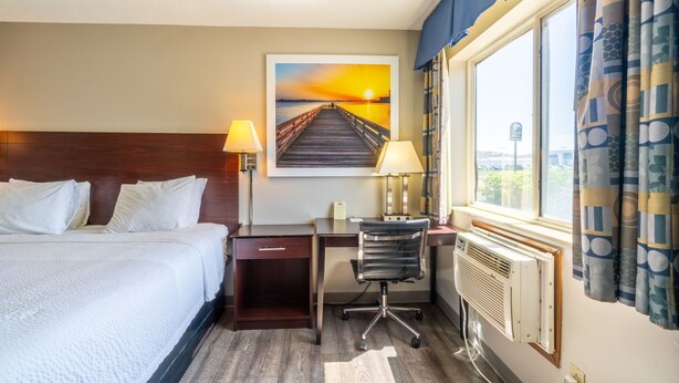 A hotel room with a bed, desk, chair, and a painting of a sunset over a pier.