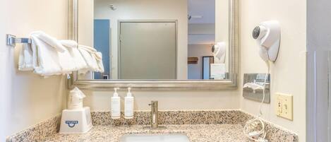 Un baño con encimera de granito, lavabo, espejo, toallero, secador de pelo y artículos de aseo.