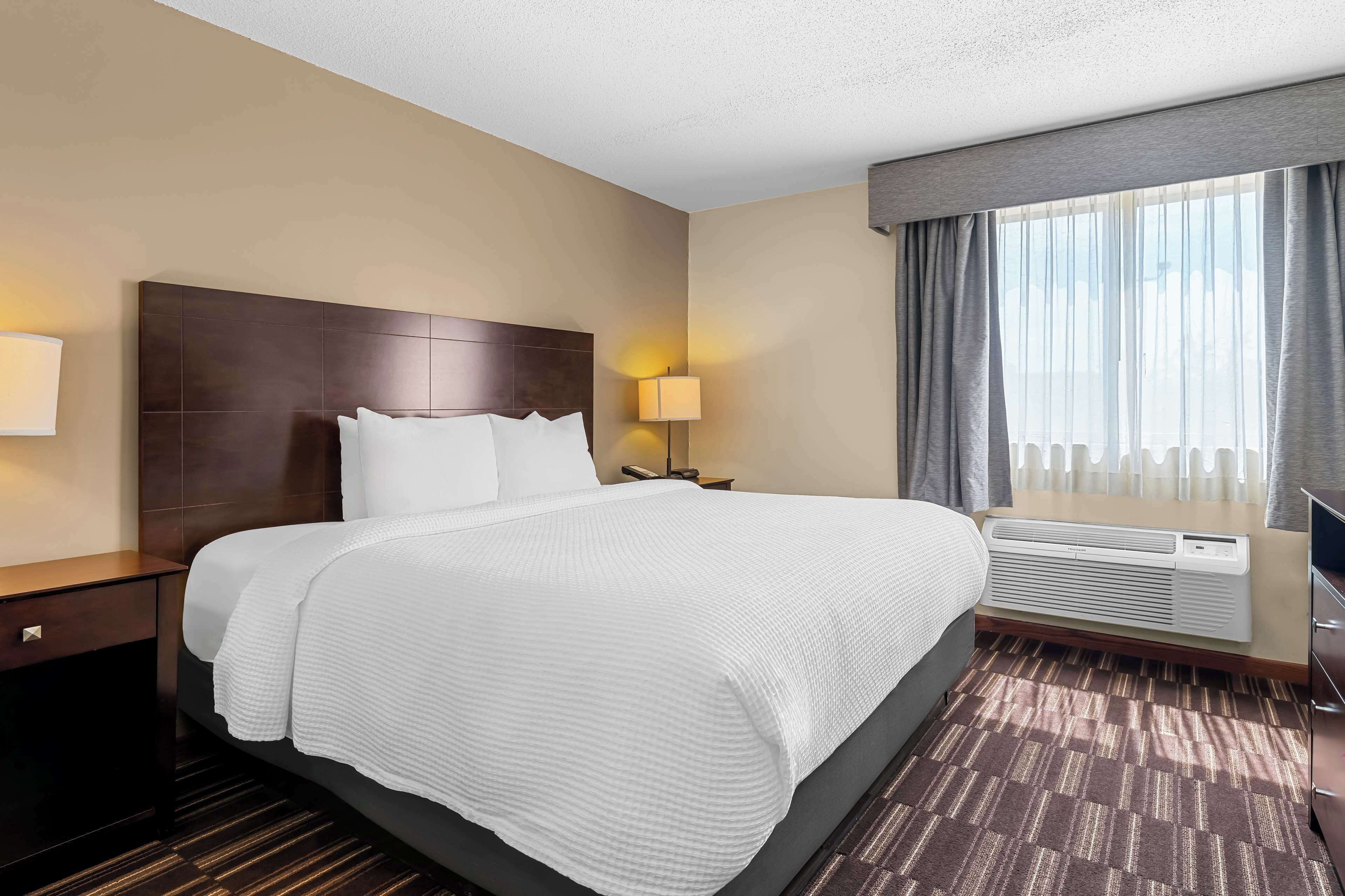 A hotel room with a large bed, a nightstand, a lamp, a window with curtains, and an air conditioning unit.