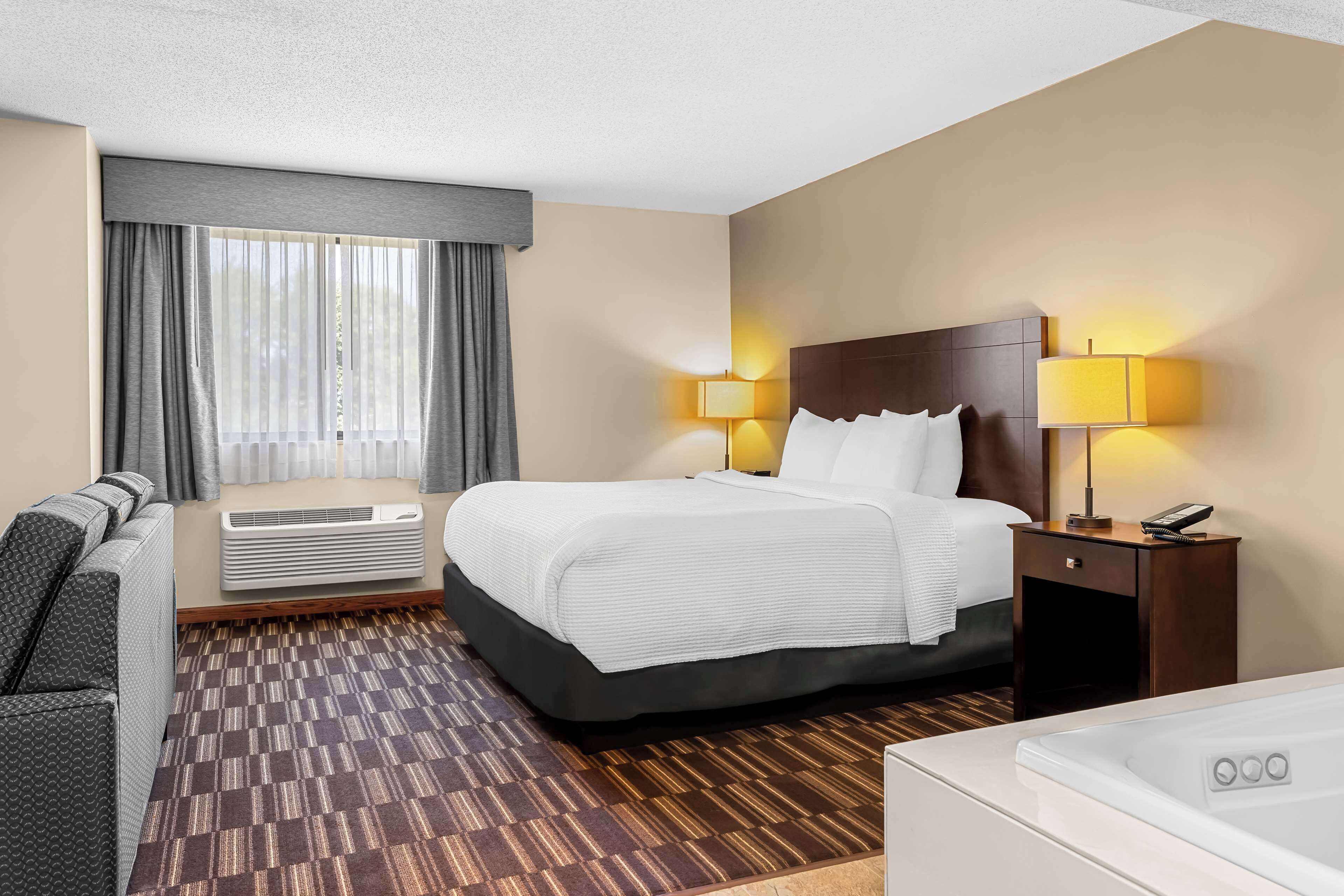 A hotel room with a bed, bedside table with a lamp, a couch, an air conditioning unit, and a window with curtains.