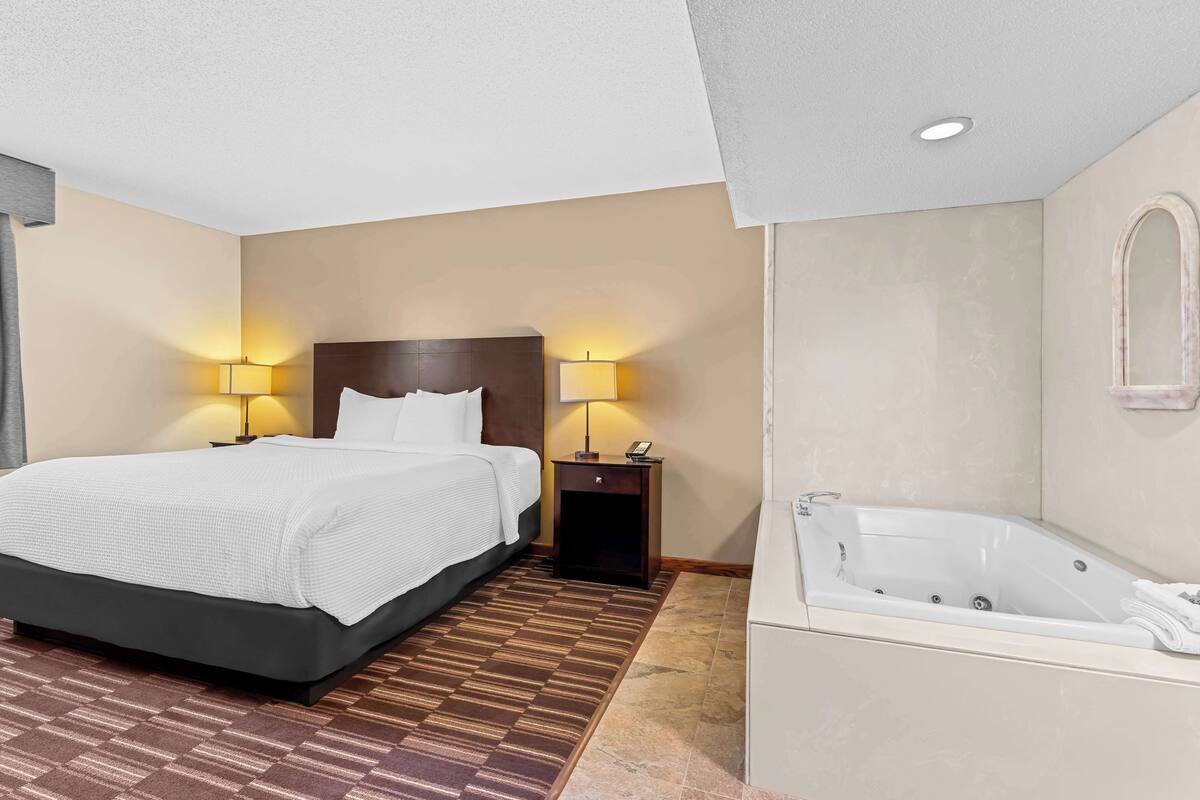 A hotel room with a large bed, two bedside lamps, a nightstand, and a separate bathroom with a bathtub.