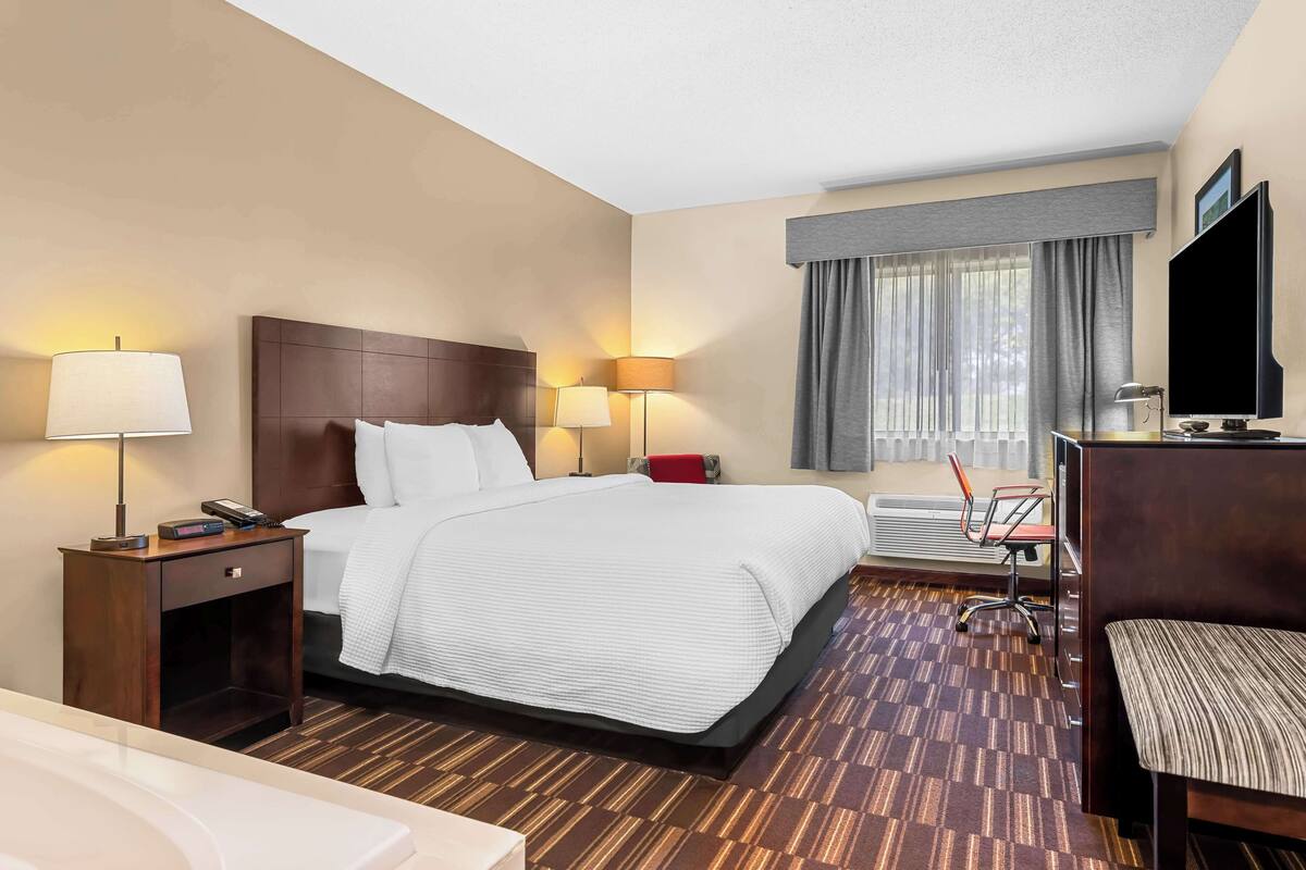 A hotel room with a large bed, two bedside tables with lamps, a desk with a chair, a TV, and a window with curtains.
