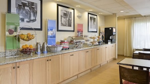 A hotel breakfast area with a buffet setup, including fruits, beverages, and a coffee machine.