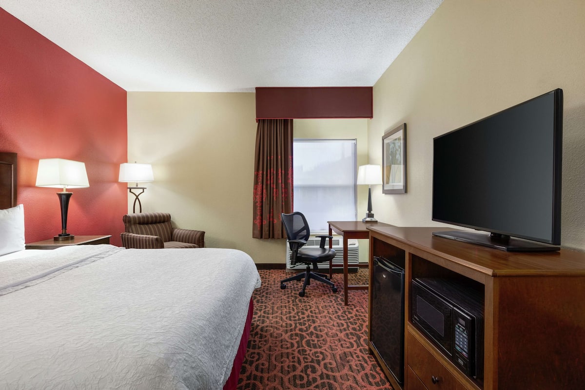 Quarto de hotel com uma cama grande, uma escrivaninha com televisão de tela plana, uma cadeira, abajur, micro-ondas e janela com cortinas.