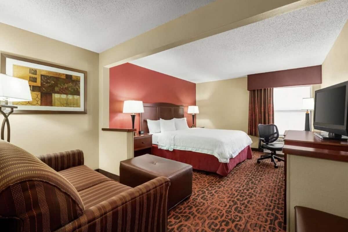 Quarto de hotel com cama, escrivaninha, computador, cadeira, abajur e televisão.