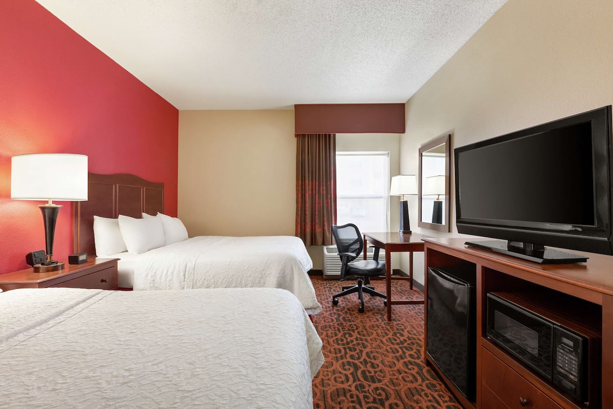 Quarto de hotel com duas camas, uma mesa de trabalho com televisão, uma cadeira e um micro-ondas.