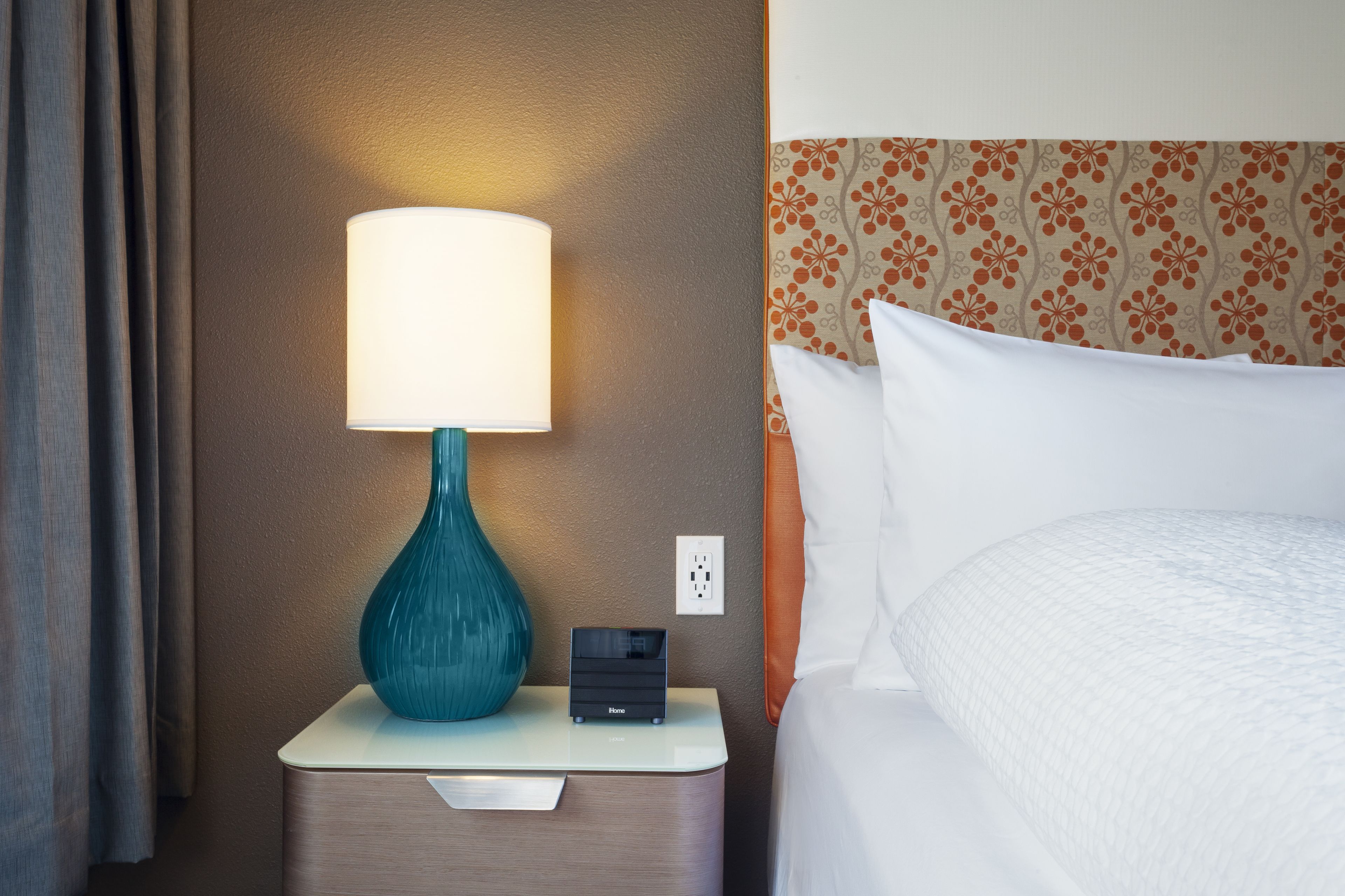 Een hotelkamer met een bedlampje, een klein nachtkastje, een stopcontact en een bed met een bloemenmotief op het hoofdeinde.