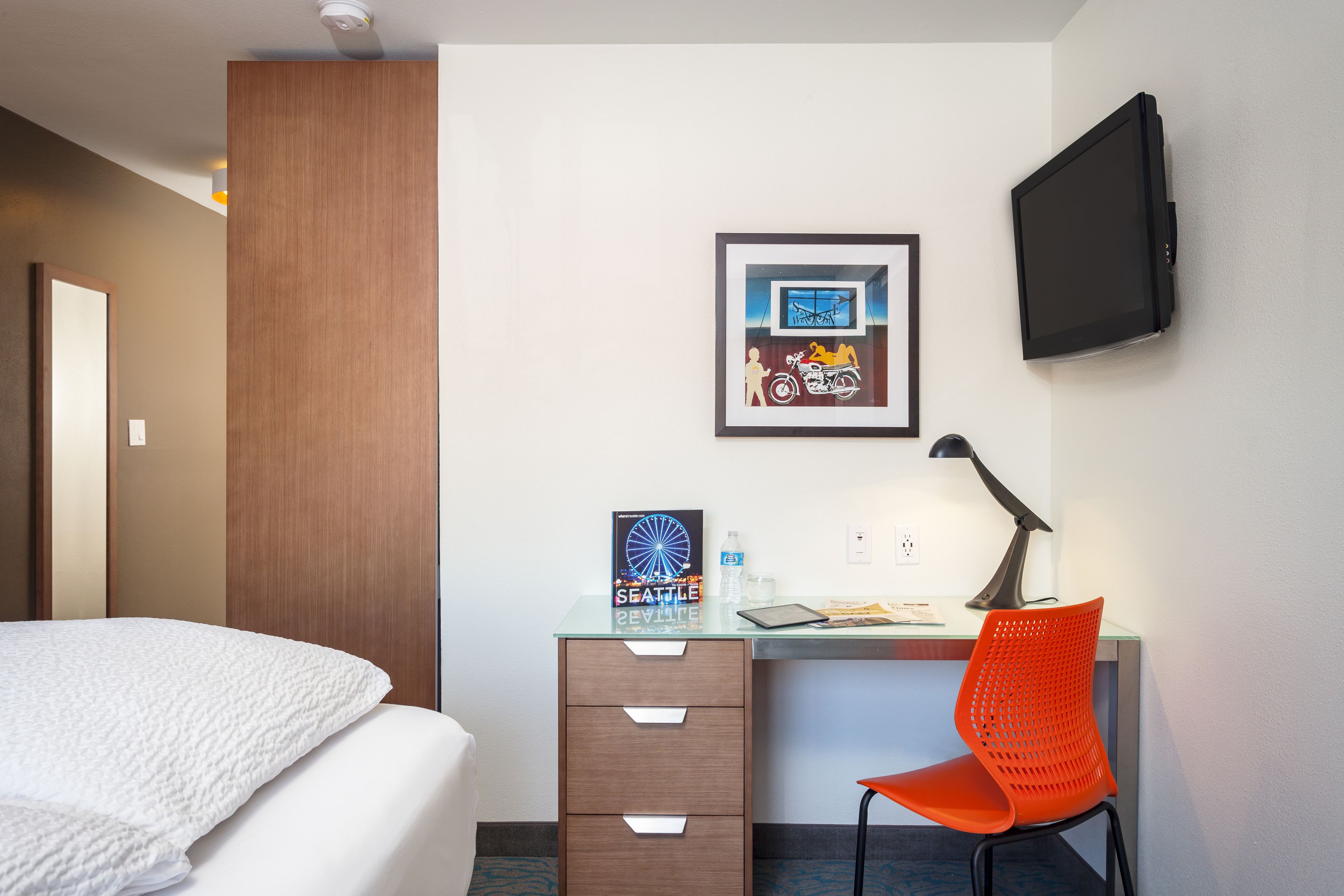 A hotel room with a bed, a desk with a chair, a television, and a framed picture on the wall.