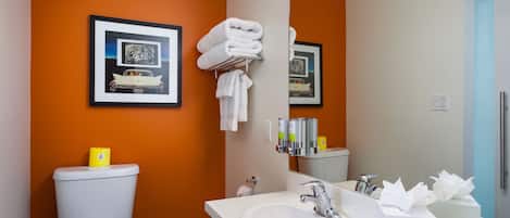 Een badkamer met een oranje wand, een witte wastafel, een toilet en lijstjes met foto’s.