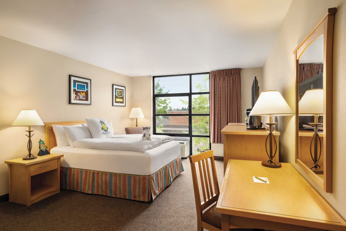 A hotel room with a bed, two bedside tables with lamps, a desk with a chair, and a large window with curtains.