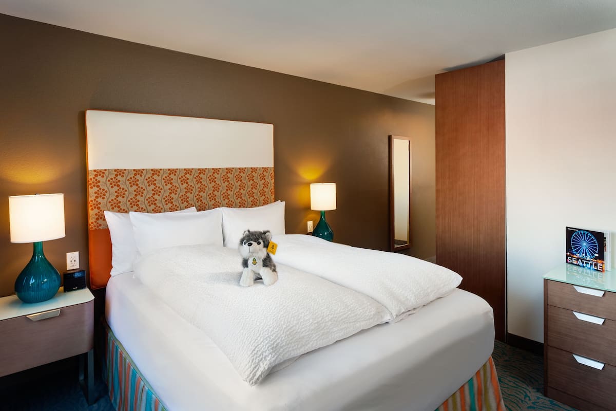 A hotel room with a large bed, two bedside lamps, a plush toy, and a framed picture on the nightstand.