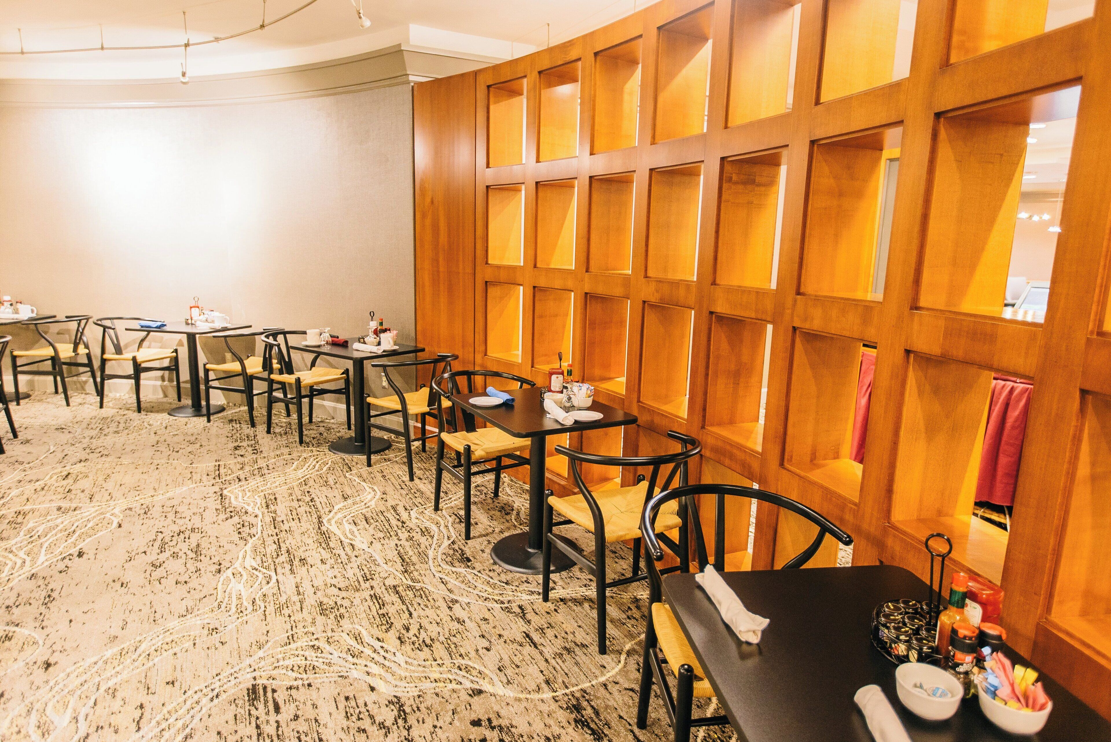 A hotel room with a dining area featuring multiple tables and chairs, a patterned carpet, and wooden paneling on the walls.