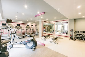Un gimnasio con cintas de correr, elípticas, una prensa de banca y un estante con mancuernas.