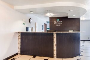 Um balcão de atendimento com acabamento em madeira escura e uma bancada de mármore. Há um relógio e uma placa acima do balcão que diz "Quality Inn & Suites". O chão tem um padrão quadriculado preto e branco.