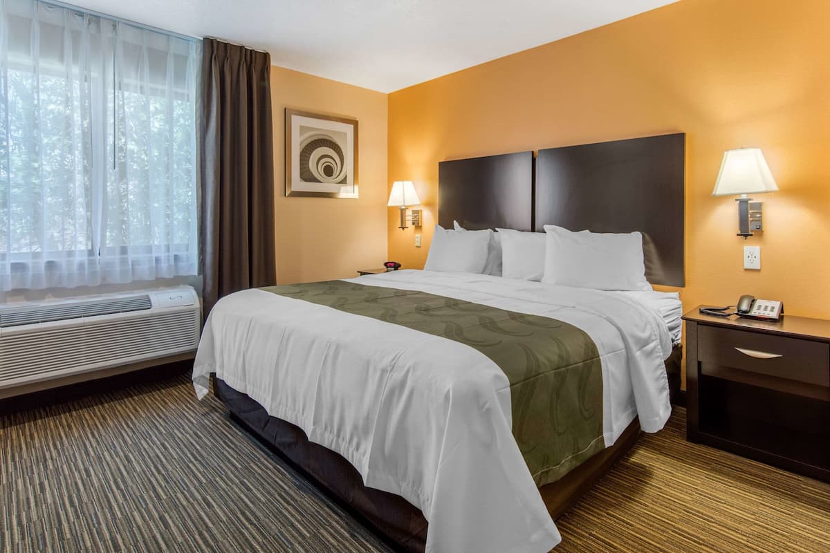 A hotel room with a large bed, two bedside lamps, a nightstand with a phone, and a window with sheer curtains.