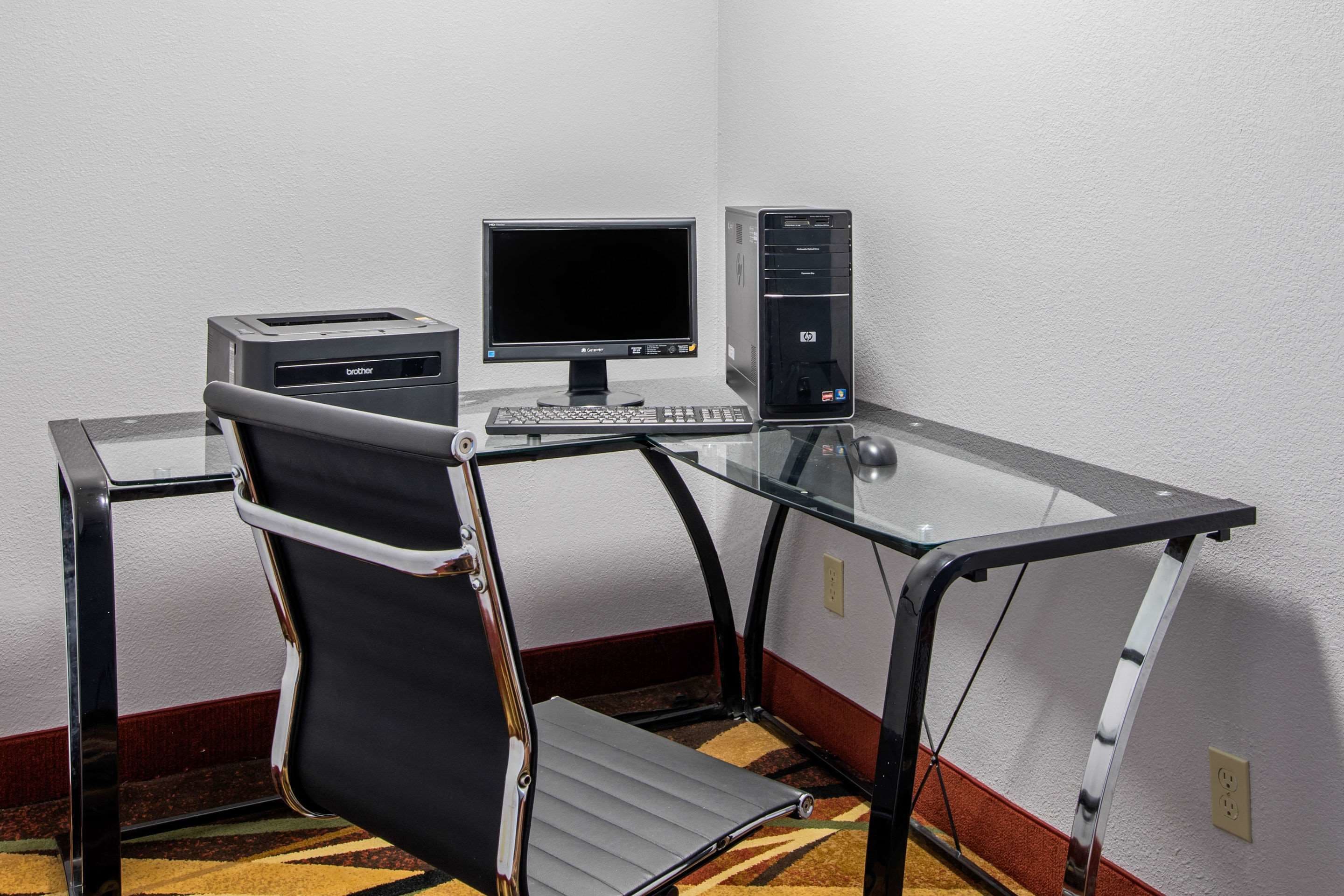 A compact hotel room with a glass desk, a black office chair, a desktop computer, and a printer.