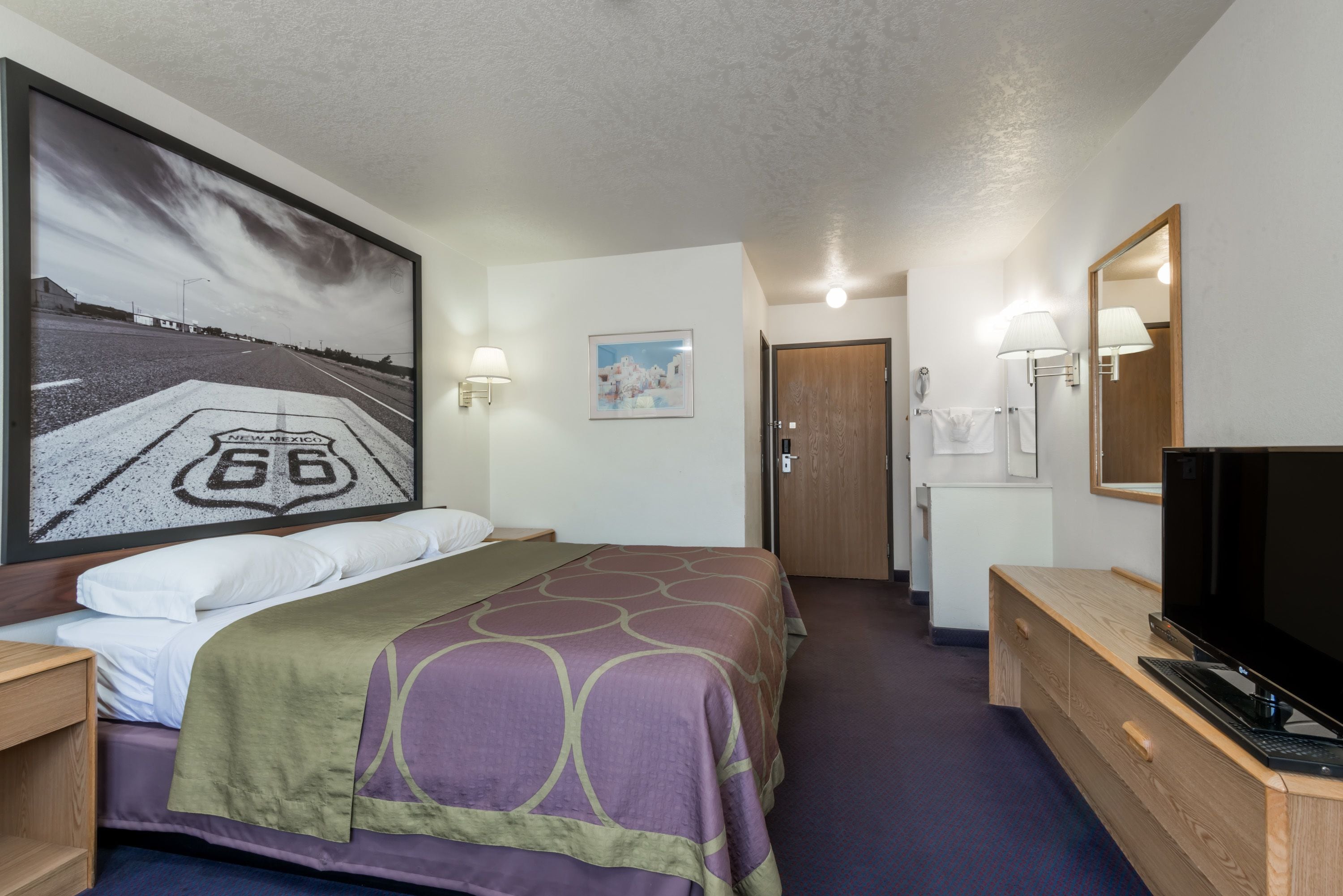 Une chambre d’hôtel avec un lit, un bureau avec une télévision, un grand tableau représentant une route, et une porte donnant sur une salle de bain.