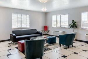 Una sala moderna con un sofá negro, sillones azules y un reposapiés rojo. Hay dos ventanas con persianas, un radiador y una planta en maceta.
