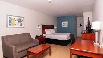 A hotel room with a brown sofa, a wooden desk, a bed with white linens, a nightstand, and a wall-mounted TV.