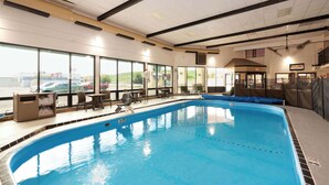 Piscina interior com paredes de vidro transparente, teto com vigas aparentes e vista para o estacionamento.