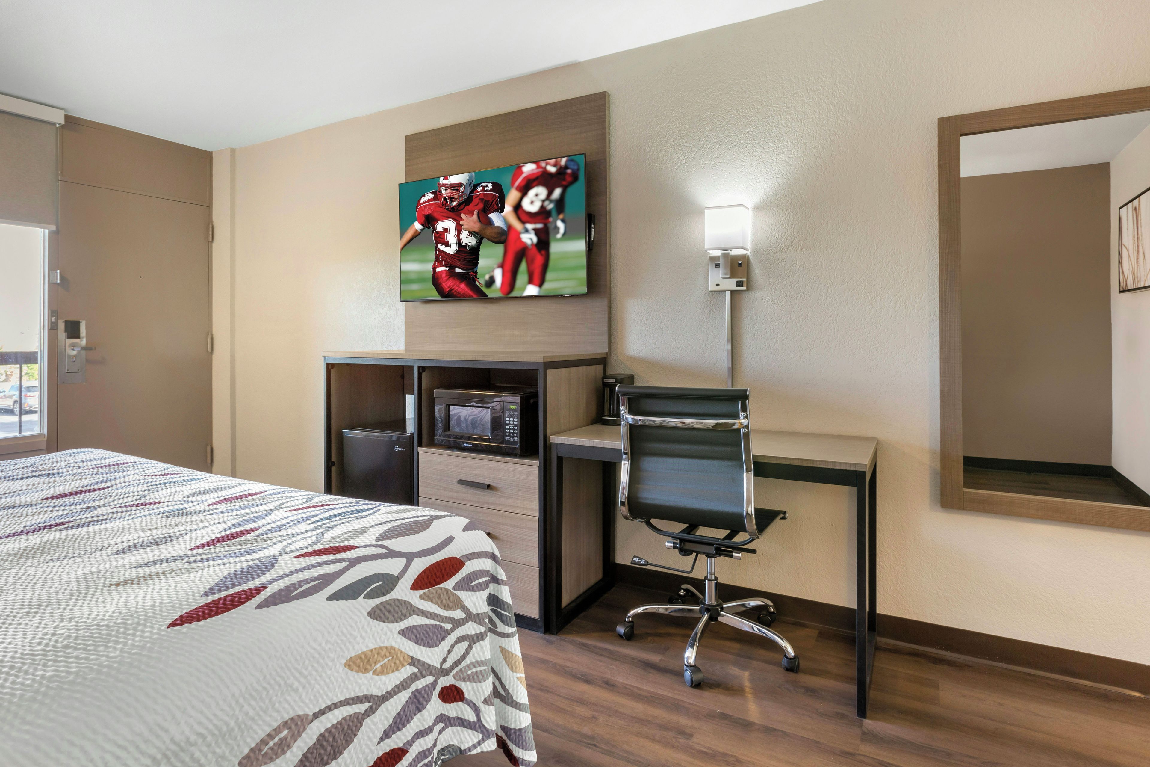 Habitación de hotel con cama, escritorio y silla, televisor, microondas y un espejo grande.