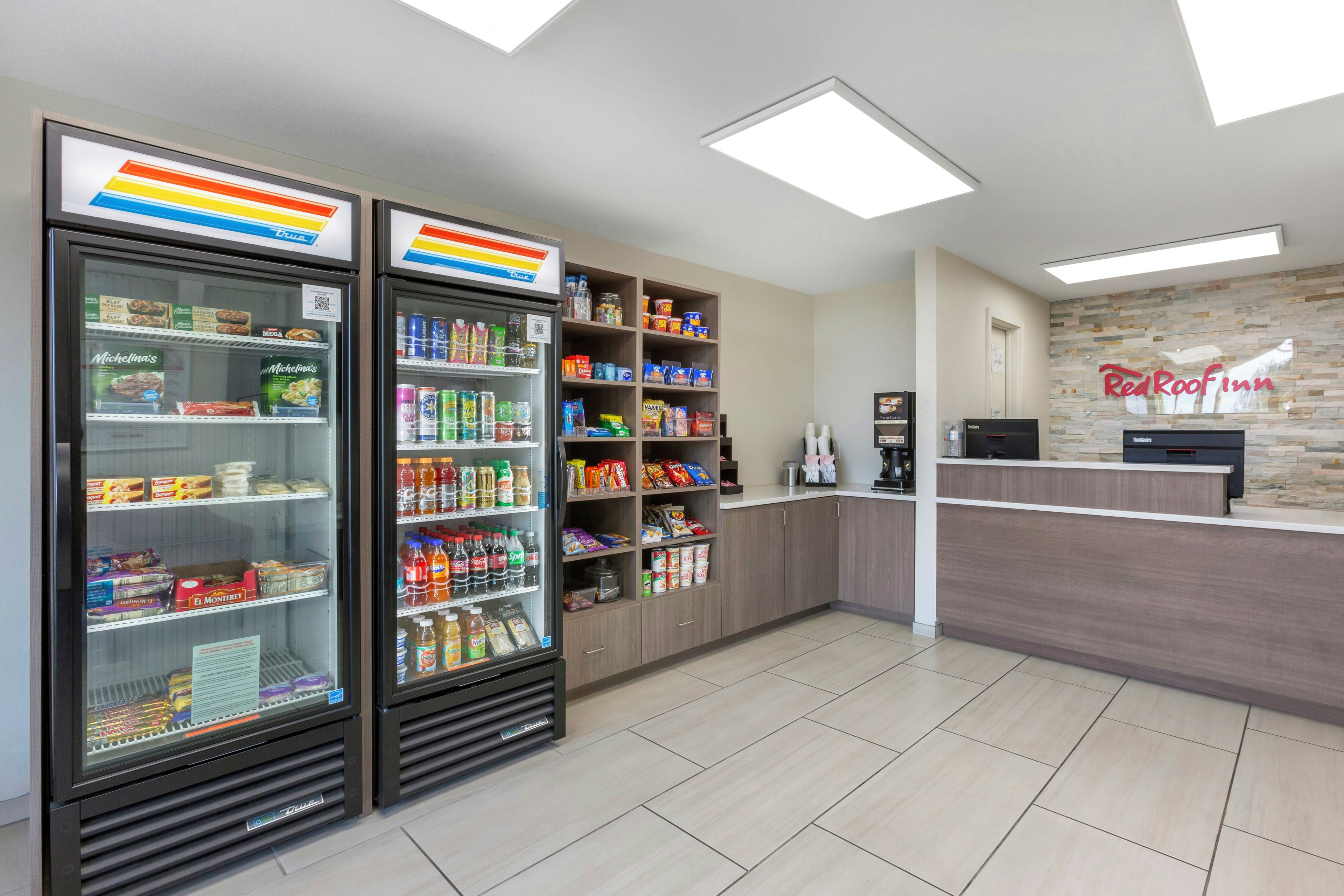 Zona de recepción de un hotel con mostrador, máquina de café y dos dispensadores refrigerados de bebidas.