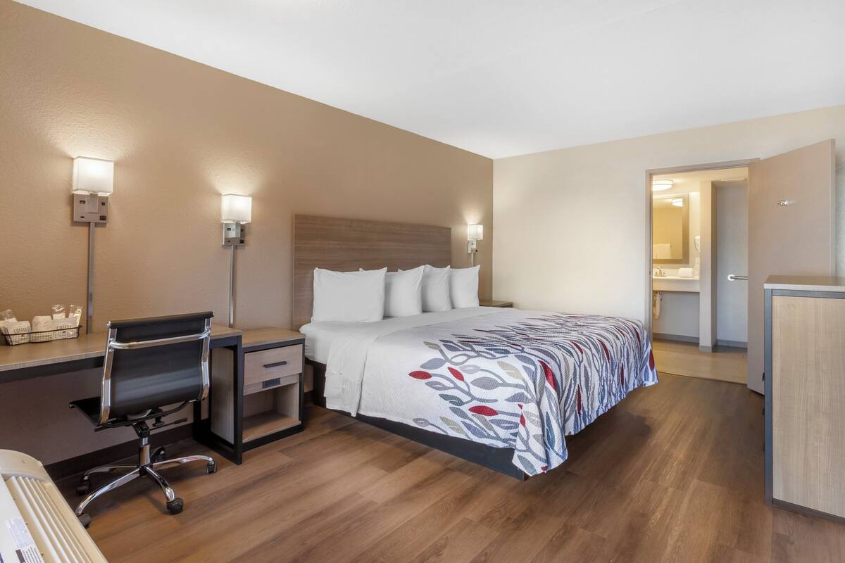 Une chambre d’hôtel avec un lit, un bureau, une chaise et une salle de bain visible à travers une porte ouverte.