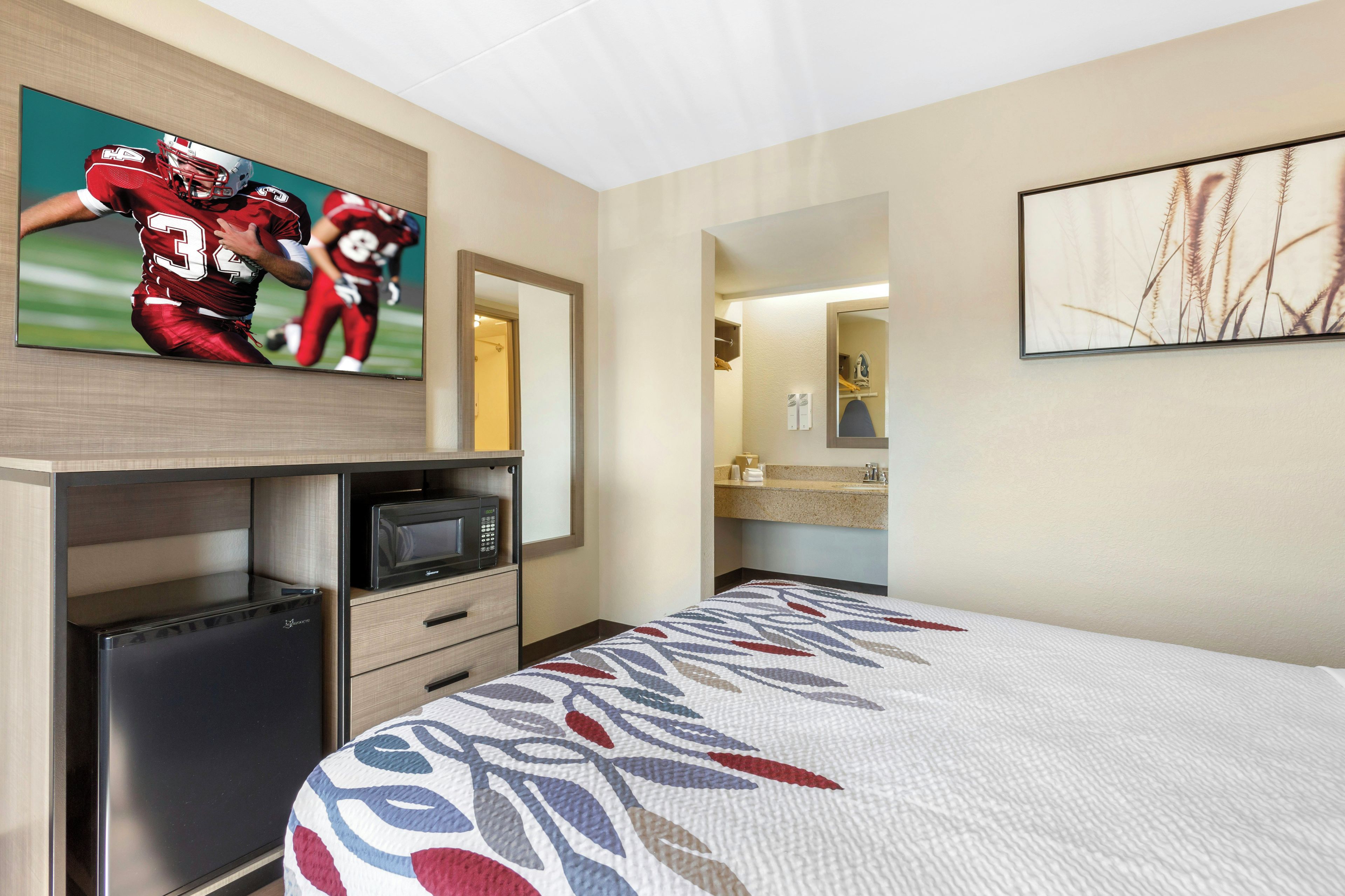 Habitación de hotel con una cama, televisor montado en la pared, microondas, refrigerador pequeño, cómoda y baño a la vista a través de una puerta abierta.