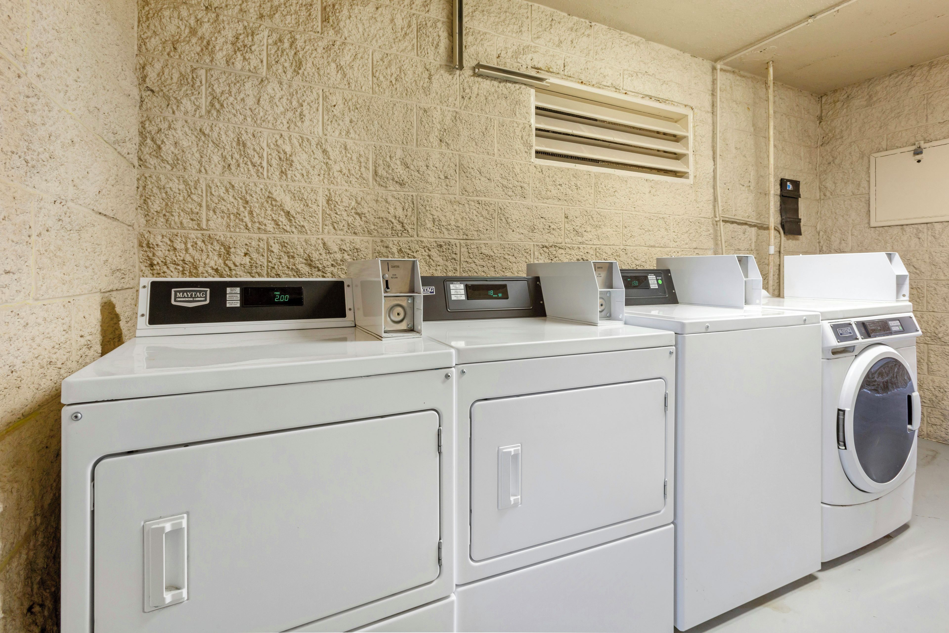 Un área de lavandería con lavadoras y secadoras apiladas, un panel de control y una ventilación en la pared.
