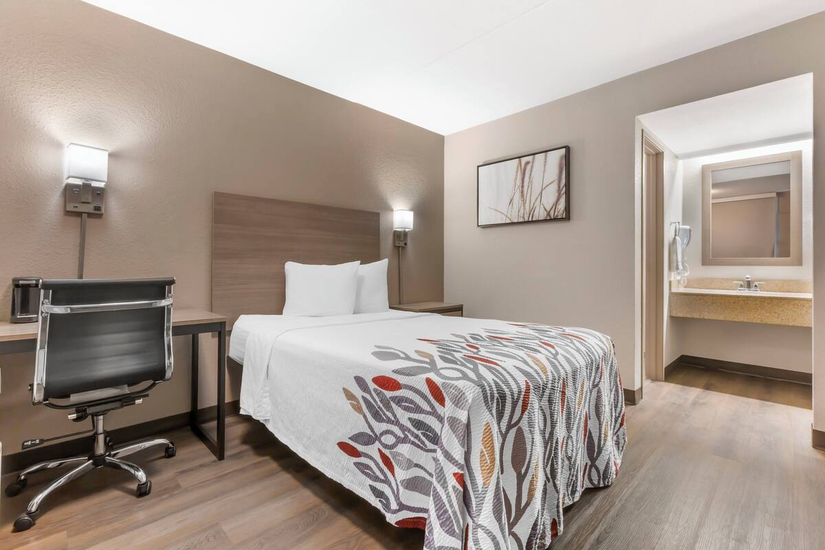 Une chambre d’hôtel comprenant un lit, un bureau avec une chaise, une table de chevet, une lampe fixée au mur et une salle de bain avec un lavabo et un miroir.