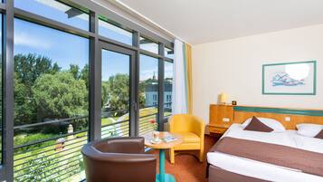 A hotel room with a large window, a bed, a chair, a small table, and a nightstand.
