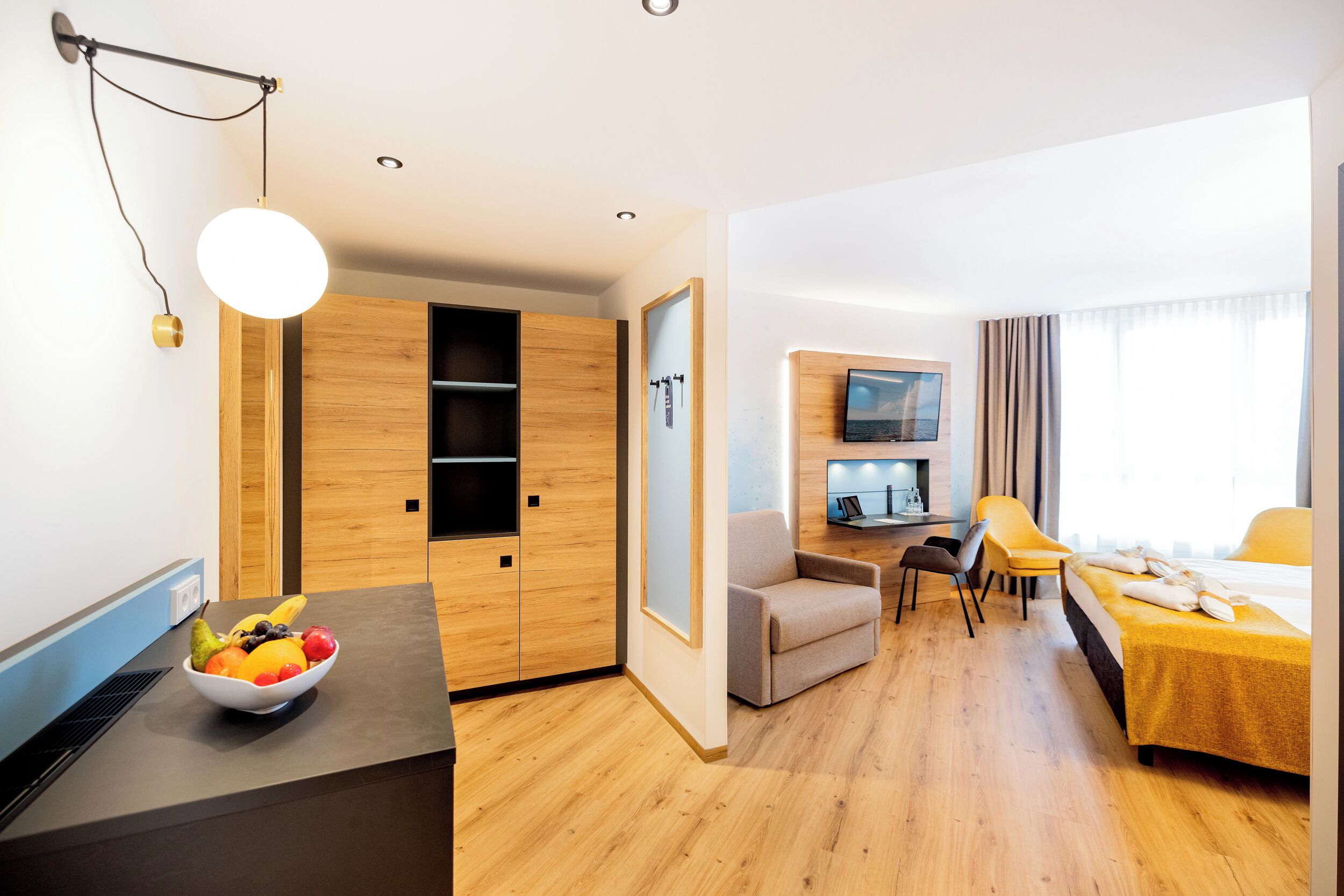 A modern hotel room with a wooden floor, a dining area with a yellow table and chairs, a bed with a yellow bedspread, a TV mounted on the wall, and a fruit bowl on the counter.
